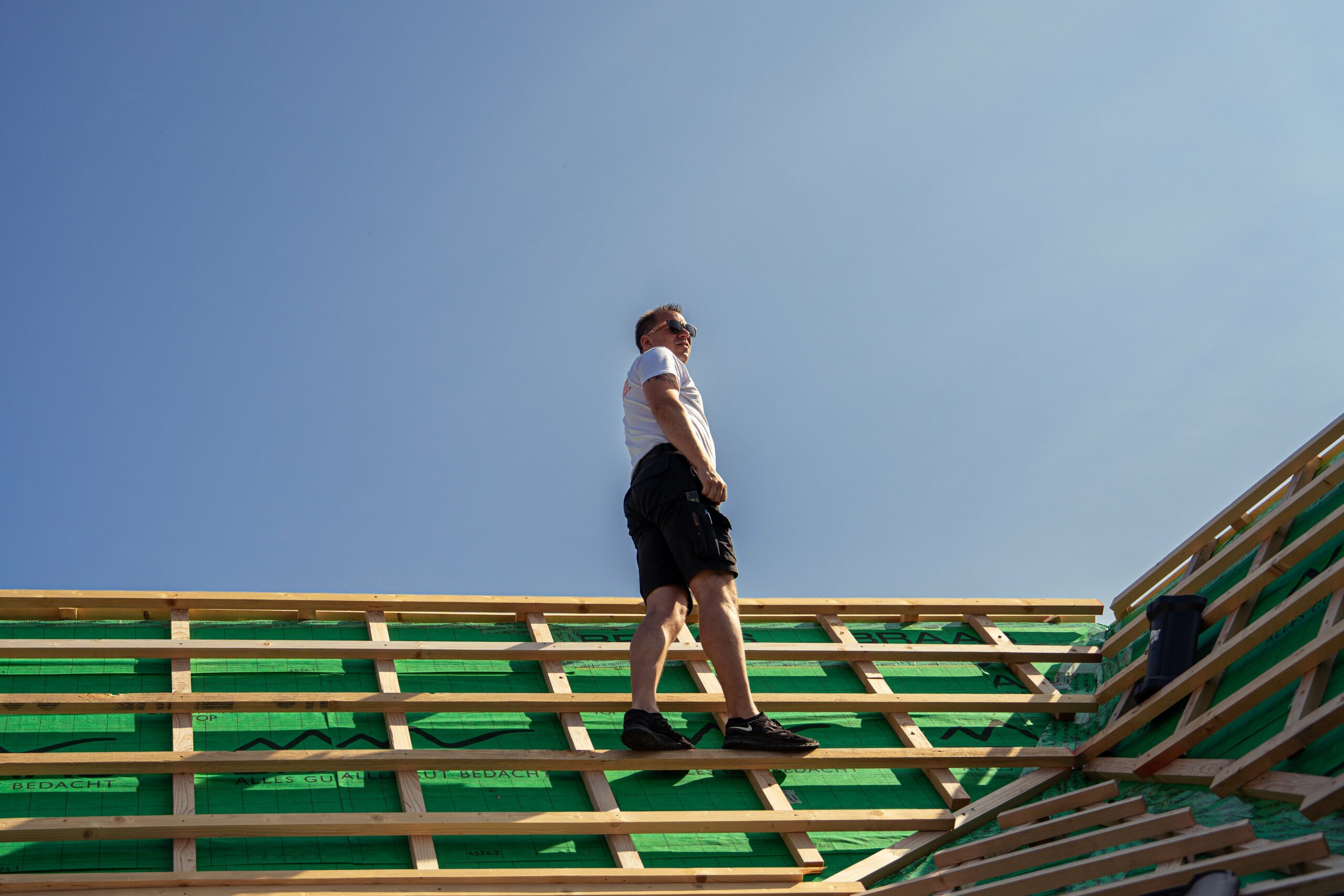 Ein Dachdecker steht auf einem Dach mit Holzrahmen und grünen Materialien unter blauem Himmel.
