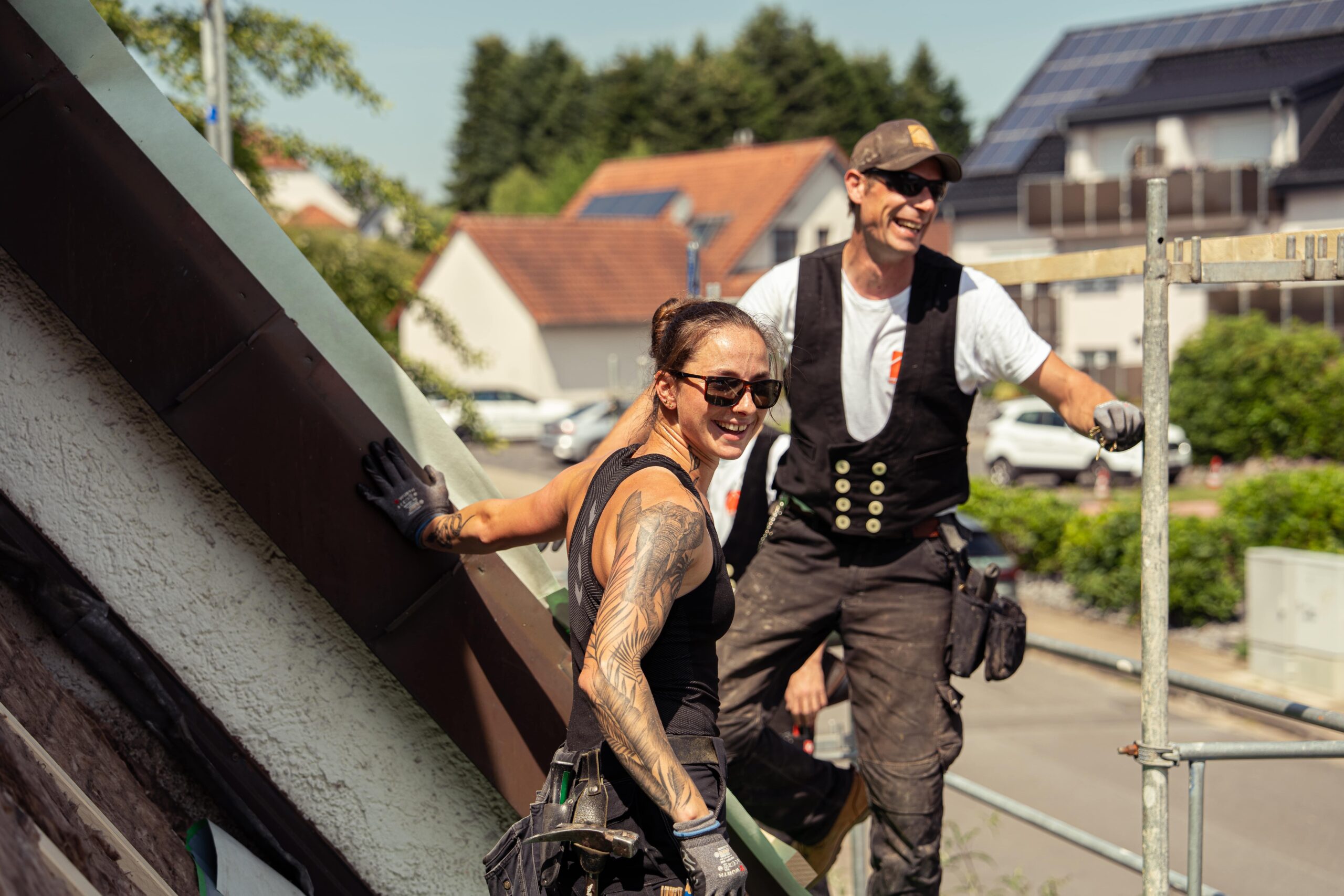 Zwei Dachdecker auf einem Dach, einer mit Sonnenbrille und tätowierten Armen, der andere lächelt.