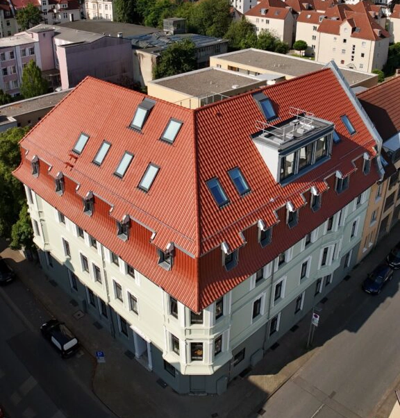 Dach eines mehrstöckigen Gebäudes mit roten Ziegeln und mehreren Dachfenstern in einer städtischen Umgebung.